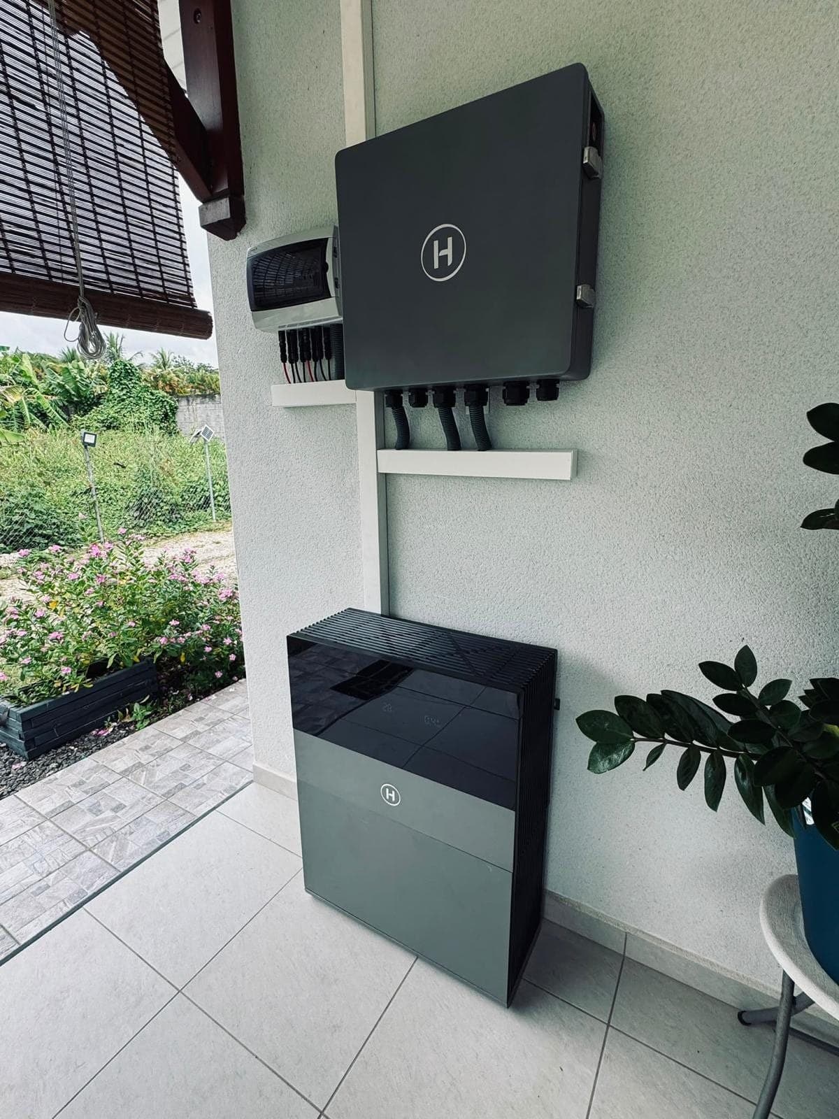 Onduleur hybride, coffret de protection et batterie fixés au mur d’une terrasse couverte, jardin en fond